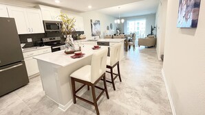 Eine moderne Küche mit weißen Schränken, einer zentralen Kochinsel mit Barhocker und einem Essbereich mit Blick zum Wohnzimmer.