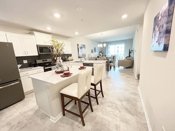 A modern kitchen with white cabinets, a central island with bar stools, and a dining area with a view to the living room.
