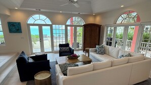 A spacious living room with a large white sofa, a coffee table, and a black armchair. There are large windows with arched tops, a ceiling fan, and a view of the ocean.