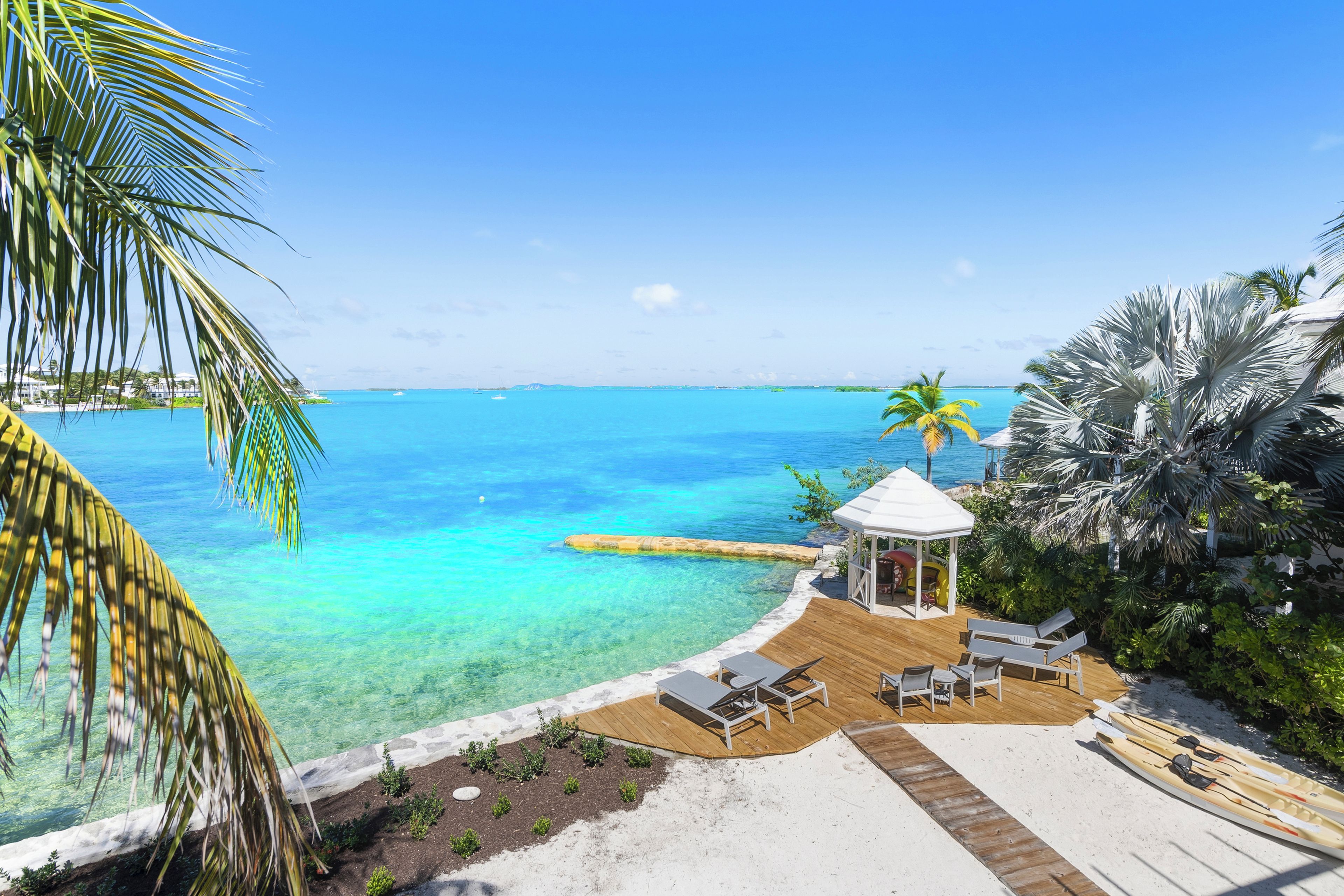 A beachside resort with clear turquoise waters, a wooden deck with lounge chairs, and a small gazebo.