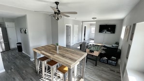 Une cuisine spacieuse avec une table en bois et des tabourets de bar, un ventilateur de plafond et une poubelle visible.