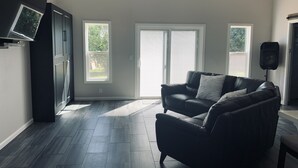 A modern living room with a dark leather sofa, a flat-screen TV mounted on the wall, and a large window with blinds.