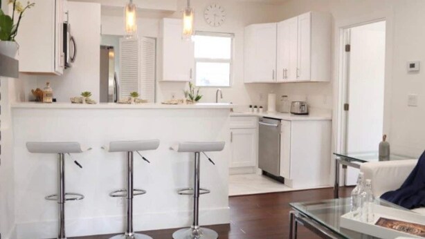 Una cocina moderna con armarios blancos, una isla central con taburetes de bar, electrodomésticos de acero inoxidable e iluminación colgante.
