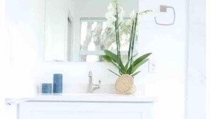 Un baño con tocador blanco, un espejo, un lavabo y un pequeño taburete de madera con una vela y una cesta tejida.