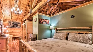 A log cabin bedroom with a wooden bed, a nightstand, a lamp, and a painting on the wall. - Cozy Log Cabin w/ Hot Tub & Fire Pit! (Sevierville)