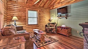 A log cabin interior with a wooden ceiling, a sofa, a chair, a table, a lamp, and a chest of drawers. - Cozy Log Cabin w/ Hot Tub & Fire Pit! (Sevierville)