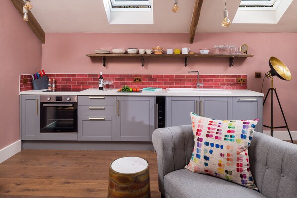 A modern kitchen with grey cabinets, a white countertop, and a pink backsplash. There is a dining area with a grey sofa and a colorful pillow. A round table is in the foreground. - The Duchy Barn at Tregoose Old Mill (Newquay)