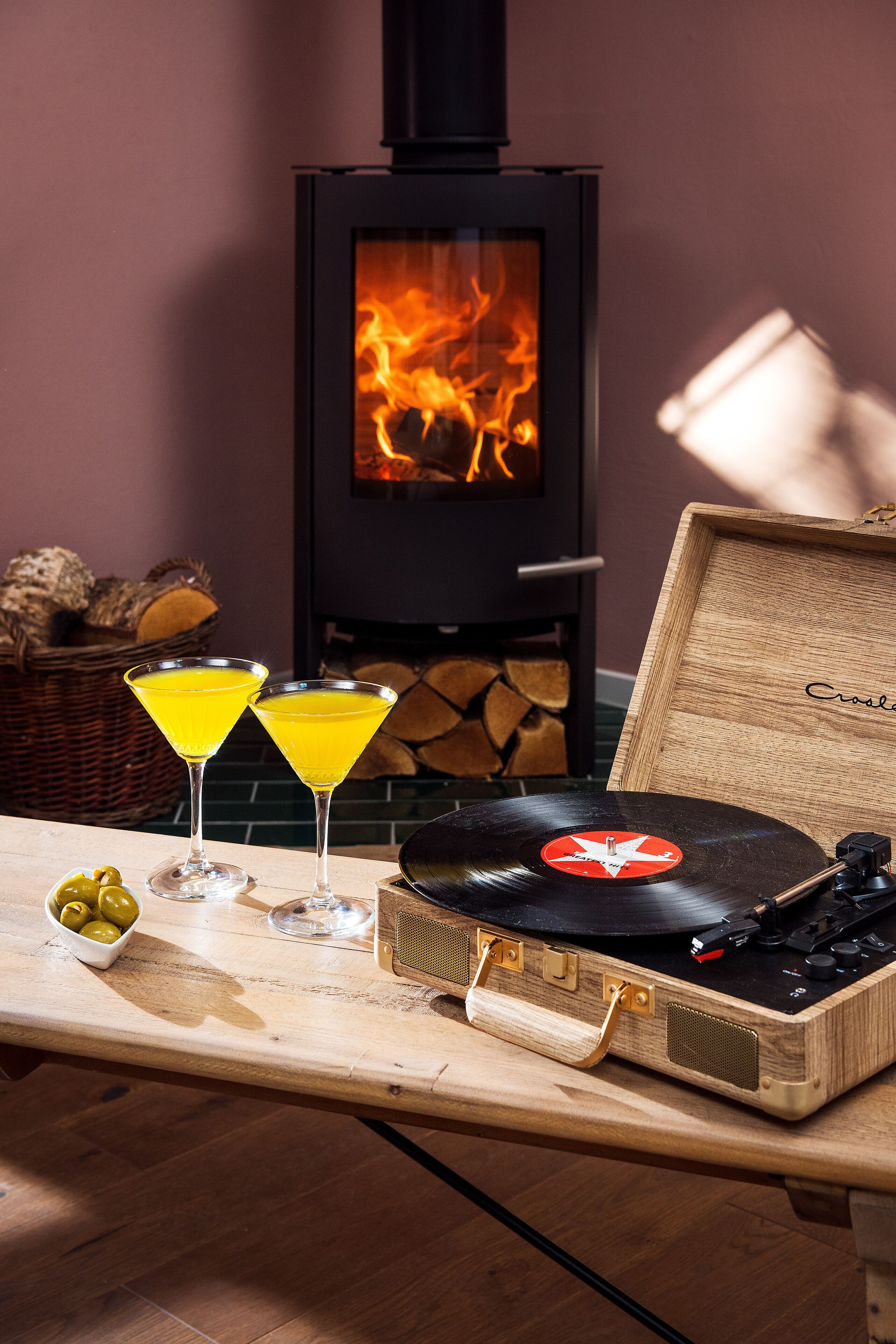Ein gemütliches Zimmer mit Kaminofen, Plattenspieler und zwei Cocktails auf einem Holztisch.