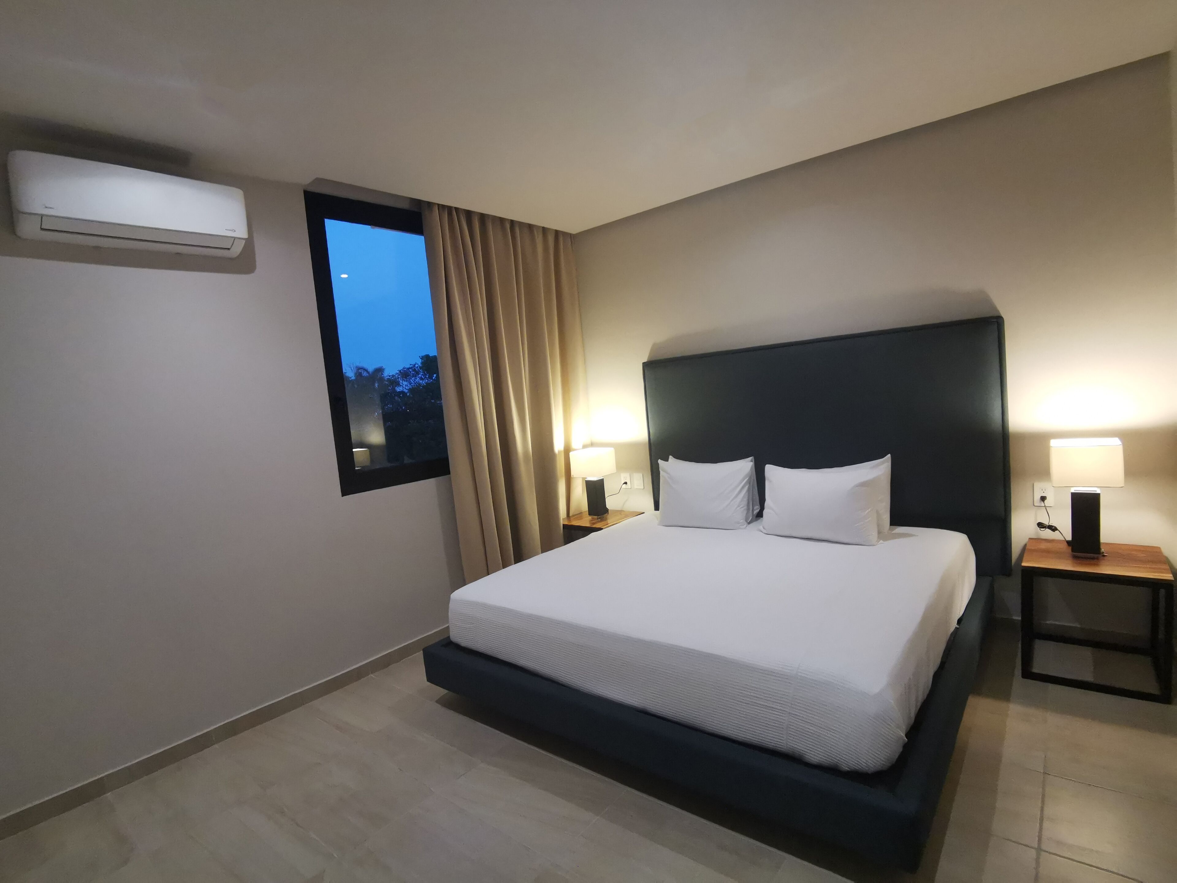 A hotel room with a large bed, two bedside tables with lamps, a window with curtains, and an air conditioning unit on the wall.