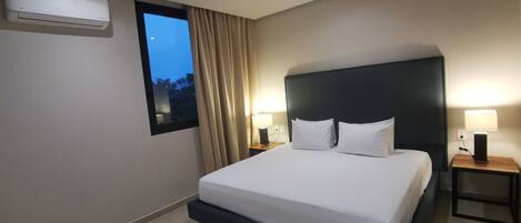 A hotel room with a large bed, two bedside tables with lamps, a window with curtains, and an air conditioning unit on the wall.