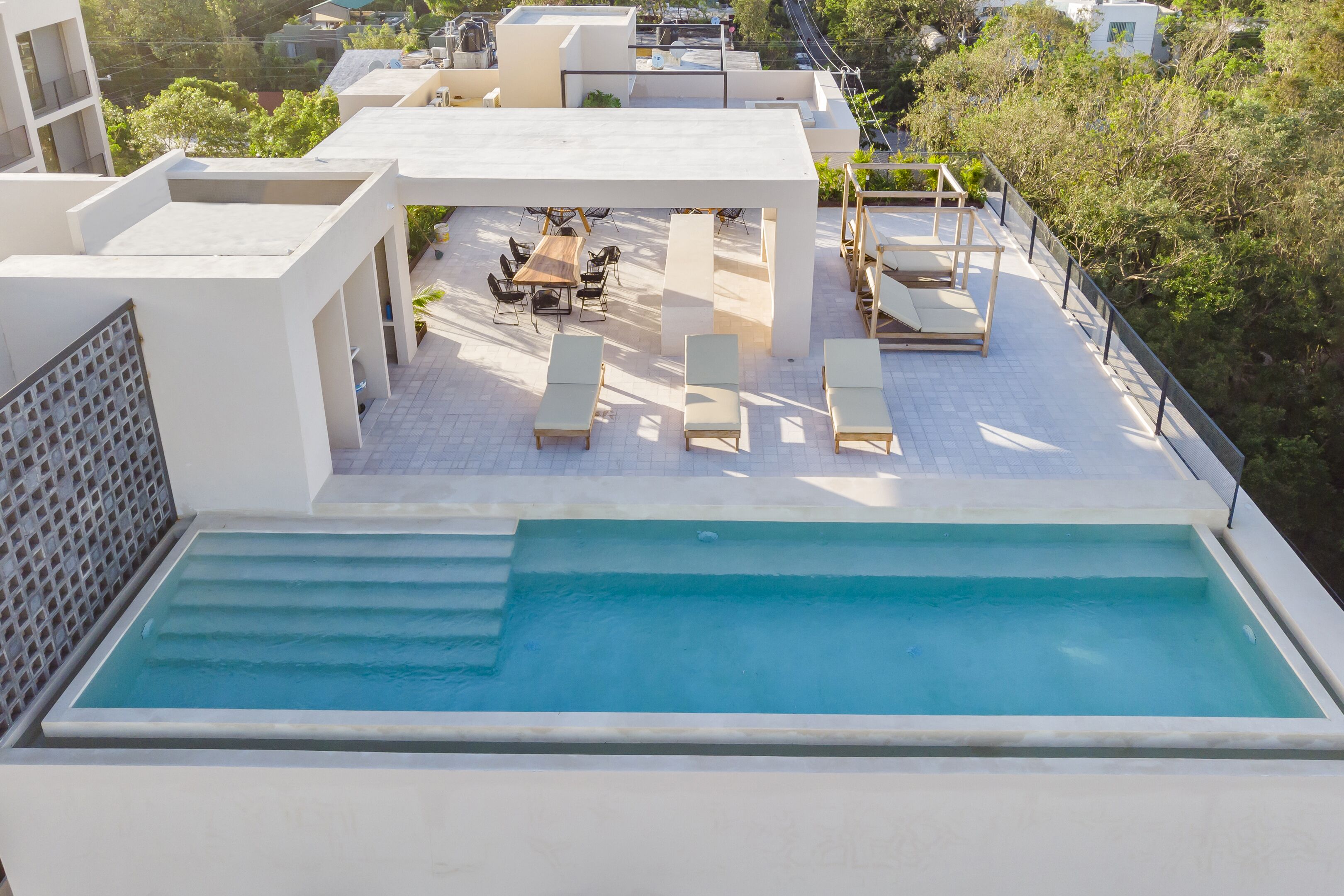 A rooftop pool area with lounge chairs, a dining table, and a clear view of the surrounding buildings and trees.