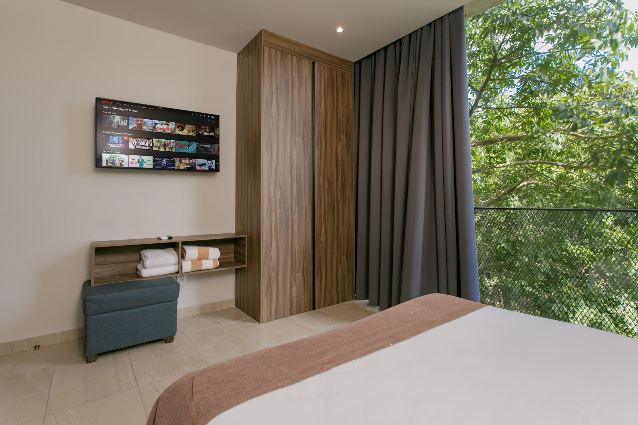 Un dormitorio moderno con una cama grande, un televisor instalado en la pared que muestra un servicio de streaming, un armario de madera, una pequeña zona de estar y un balcón con vistas a árboles.