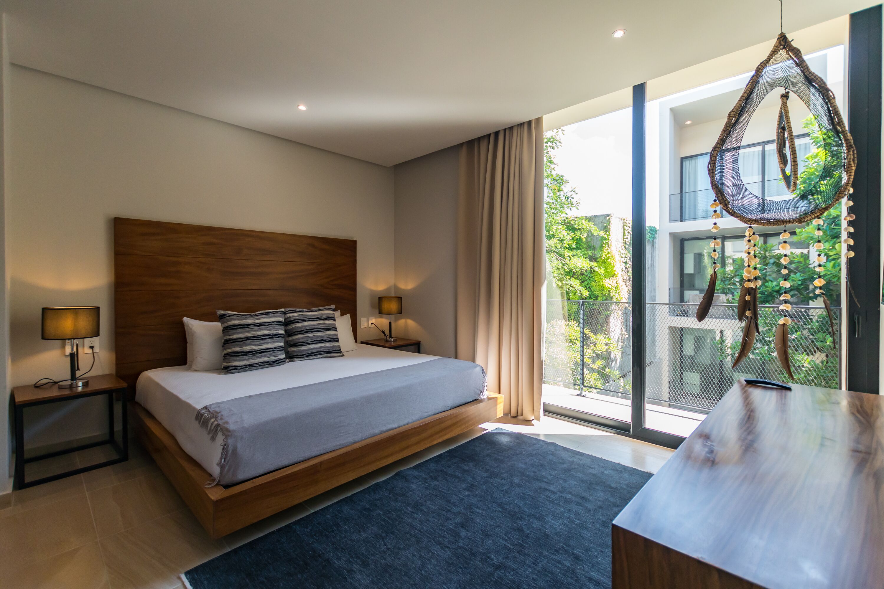 A modern bedroom with a large bed, bedside lamps, a view of greenery through large windows, and a balcony with a hanging chair.