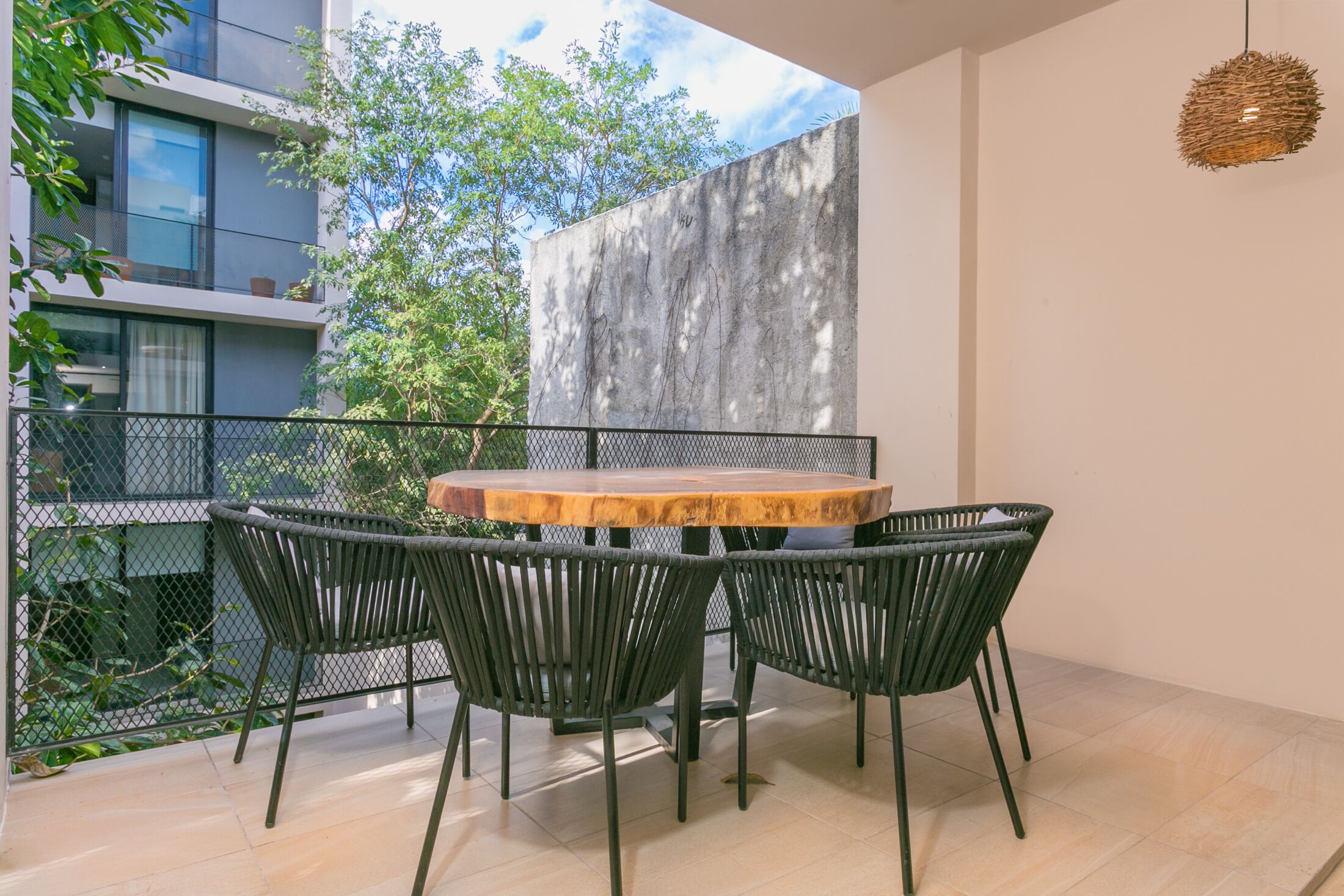 A modern outdoor dining area with a round wooden table and four black chairs, surrounded by a metal fence and greenery.