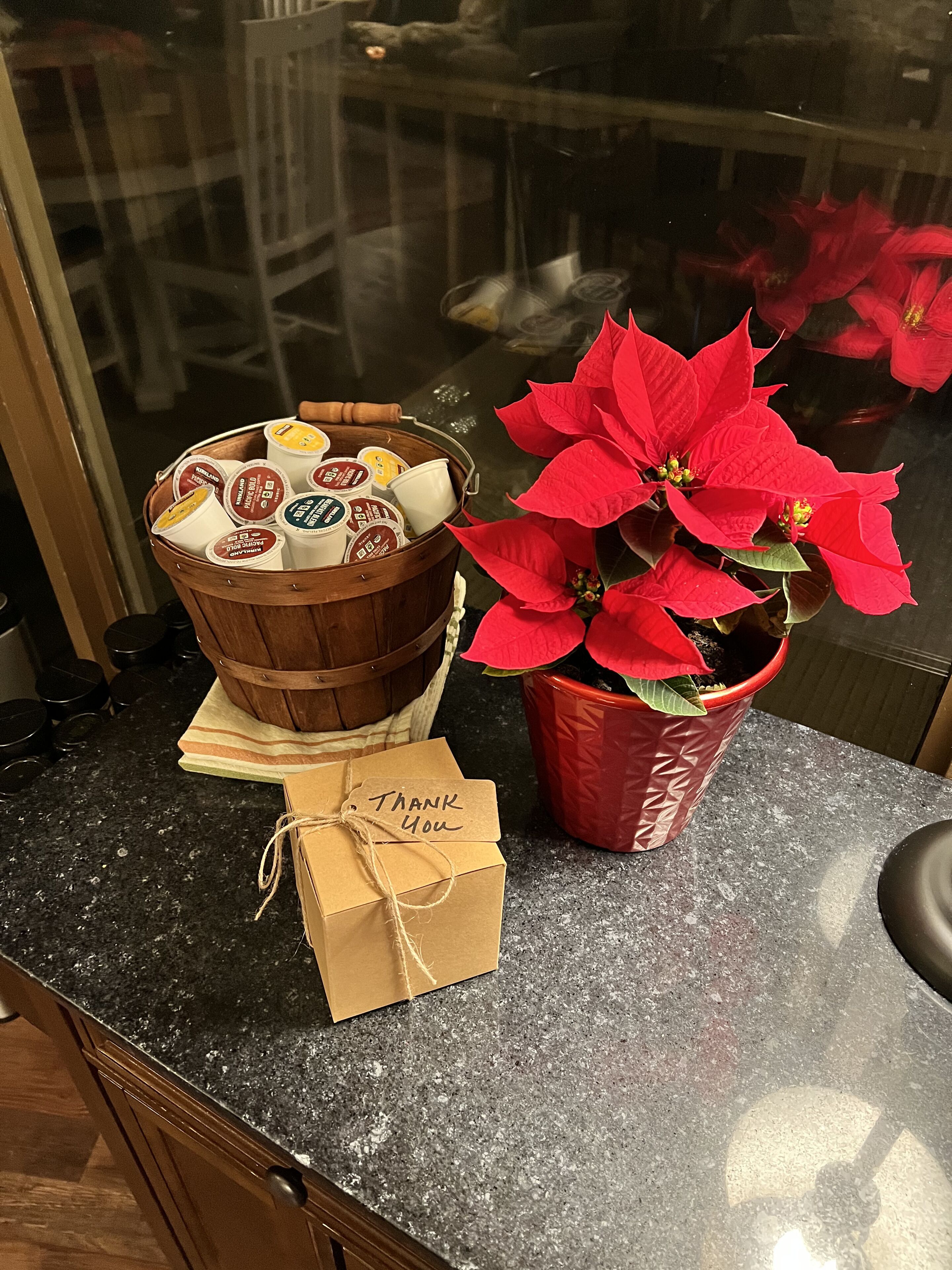Una encimera de cocina con una cesta de madera que contiene vasos de yogur, una planta de buganvilla en maceta y una pequeña bolsa de papel marrón con el texto 
