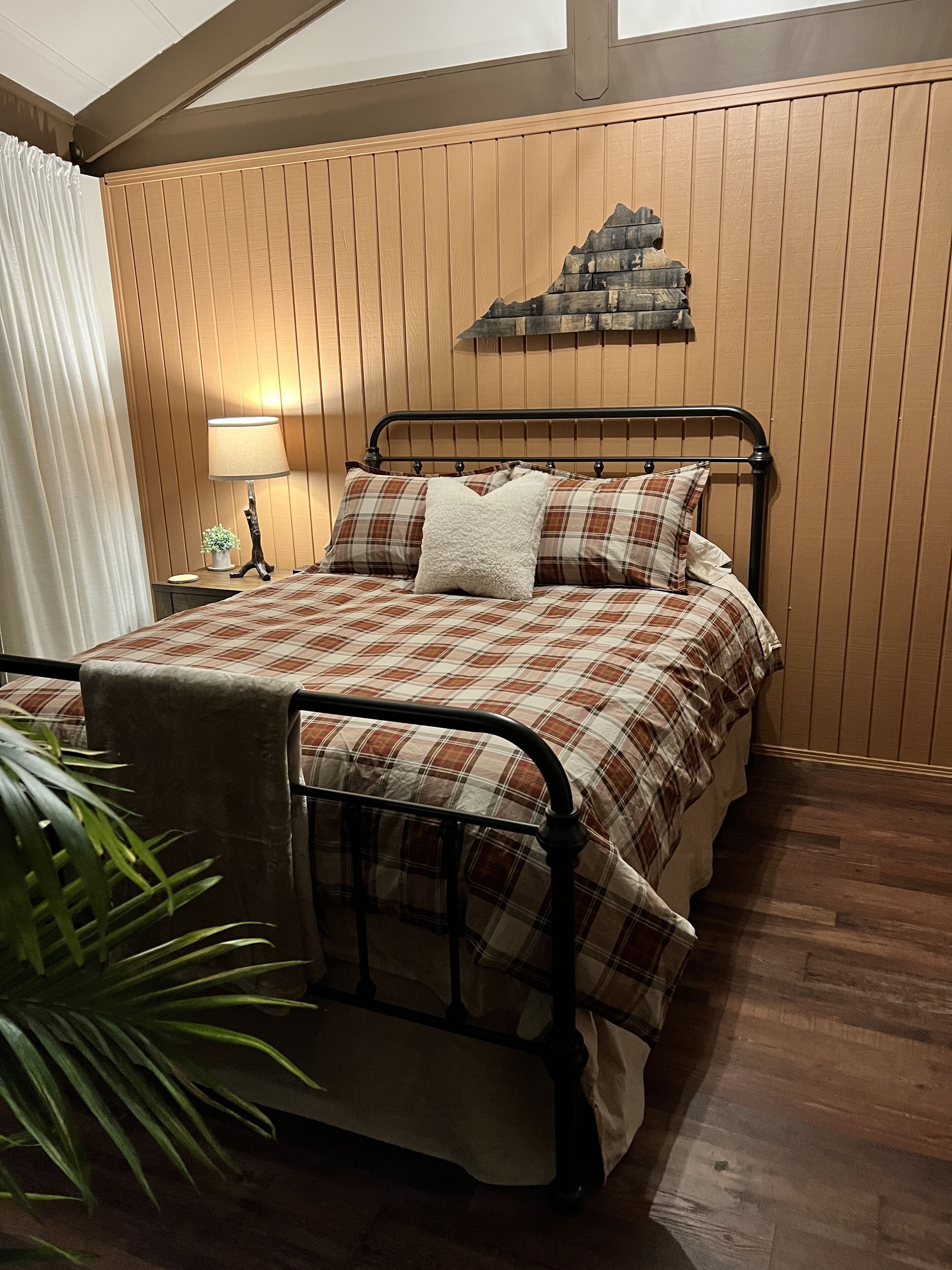 Une chambre à coucher avec un lit en métal, une couette à carreaux, une tête de lit en bois, une table de chevet avec une lampe et une plante en pot.