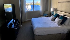 A hotel room with a large bed, a TV, a chair, and a view of the water and buildings. - 5 Stars Apart-Suites by Hilton. Amazing views. (Fort Lauderdale)