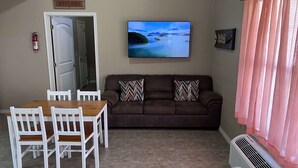 Un salon avec un canapé marron, deux coussins décoratifs à motifs, une table à manger en bois avec quatre chaises blanches, un téléviseur à écran plat fixé au mur et un extincteur également fixé au mur.