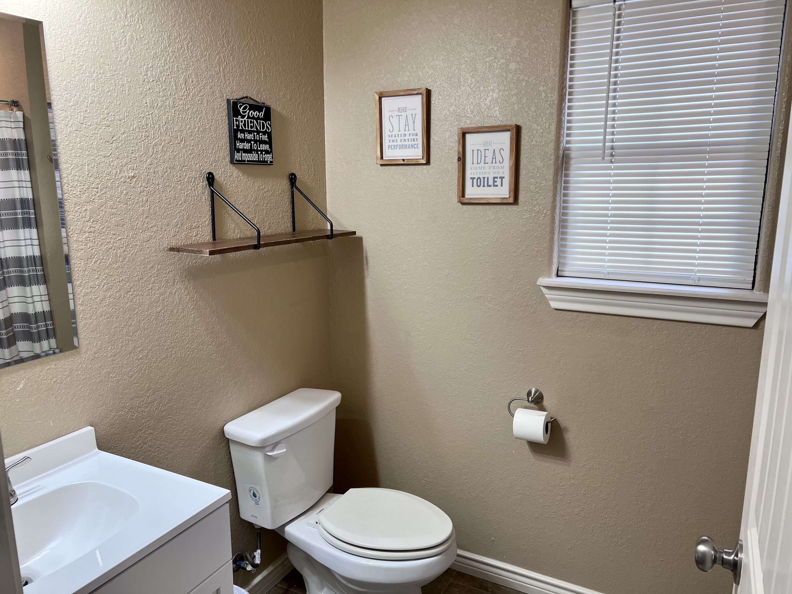 A bathroom with a toilet, sink, and a window with blinds. There are three wall-mounted signs with quotes.