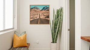 Een hal met een lijstfoto van een woegend landschap, een bankje met een geel kussen en een hoge groene plant.