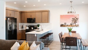 Een moderne keuken met houten kastjes, een zwarte koelkast, een magnetron en een fornuis. Er is een zithoek met een houten tafel en stoelen, en een bank met kussens.