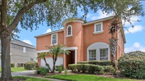 A two-story house with a red exterior, white trim, and a garage. It has a well-maintained lawn and several palm trees. - Be-Our-Guest Home! 7 bdrm incl. 5 King beds, Private POOL, 15 minutes to Disney! (Kissimmee)