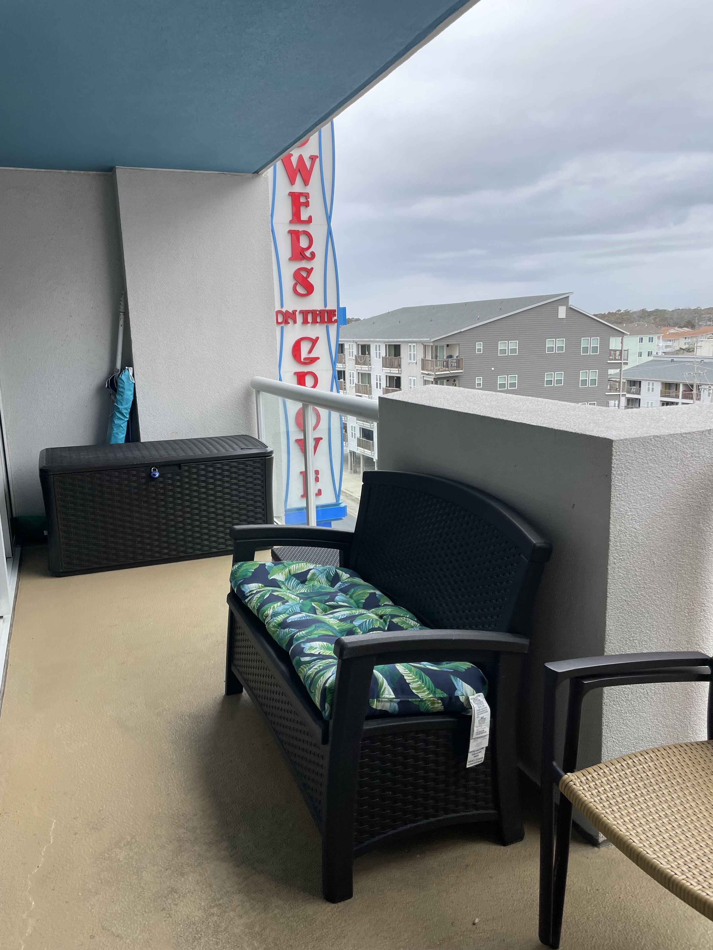 A balcony with a wicker sofa and chair, a blue and white banner, and a view of buildings in the background.