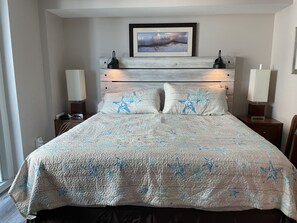 Cama bem arrumada com colcha estampada em formato de estrela, duas luminárias de cabeceira, um quadro acima da cabeceira e uma mesinha de cabeceira pequena com uma luminária.