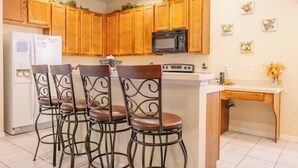 Une cuisine avec des meubles de rangement en bois, un réfrigérateur blanc, un micro-ondes noir et un tabouret de bar avec une structure en métal et un siège en cuir.