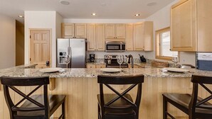 A modern kitchen with wooden cabinets, a granite countertop, and stainless steel appliances. There are three bar stools and a dining table.