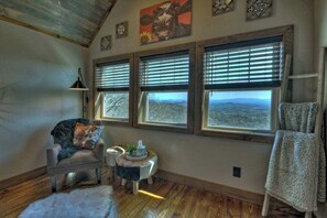 A cozy room with a leather chair, a small round table with a lamp, and a view of the mountains through large windows.
