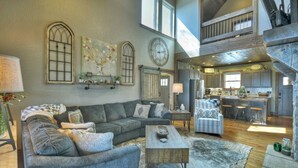 A spacious living area with a large sofa, a coffee table, and a rug. There is a kitchen area with a bar and stools. - Tranquility Perch | 3-State Mtn Views + Hot Tub (Murphy)