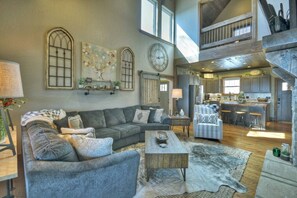 A spacious living area with a large sofa, a coffee table, and a rug. There is a kitchen area with a bar and stools. - Tranquility Perch | 3-State Mtn Views + Hot Tub (Murphy)