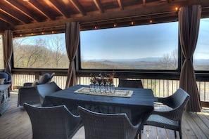 Un espace extérieur aménagé, abrité et doté de mobilier en osier, d’une table ornée d’un vase de fleurs et offrant une vue sur le paysage grâce à de grandes fenêtres.