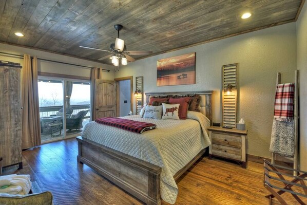 A bedroom with a large bed, wooden flooring, a ceiling fan, and a view through a sliding glass door.