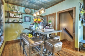 A rustic interior with a wooden dining table, two leather stools, and a hanging light fixture. - Tranquility Perch | 3-State Mtn Views + Hot Tub (Murphy)
