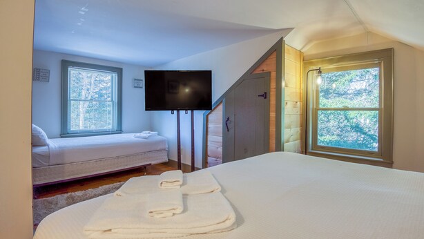 A bedroom with a bed, a TV mounted on the wall, a window with blinds, and a door leading to a loft area. - Relax-Inn - Enjoy this peaceful property along Mingo Loop (Rangeley)