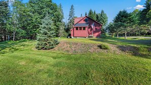 A red wooden house surrounded by a lush green landscape with tall trees. - Relax-Inn - Enjoy this peaceful property along Mingo Loop (Rangeley)