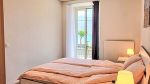 A bedroom with a bed, a nightstand, a lamp, and a view of the sea through a window.