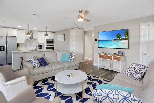 A modern living room with a sofa, coffee table, and a television displaying a beach scene. - Beachview, Fire Pit, Fenced Yard - Anchored Inn 3 (St. Joe Beach)
