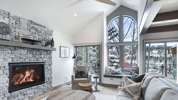 A living room with a stone fireplace, a large window with a view of trees, a sofa, and a round wooden table. - 1 Block from Main Street, Free Shuttle, Walk To Gondola, Nightlife, Shopping (Breckenridge)