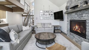 A spacious living room with a stone fireplace, a grey sofa, a round coffee table, and a staircase leading to an upper level. - 1 Block from Main Street, Free Shuttle, Walk To Gondola, Nightlife, Shopping (Breckenridge)