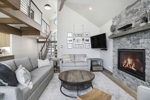 Ein geräumiges Wohnzimmer mit Kamin aus Stein, einem grauen Sofa, einem runden Couchtisch und einer Treppe, die zu einer oberen Ebene führt.