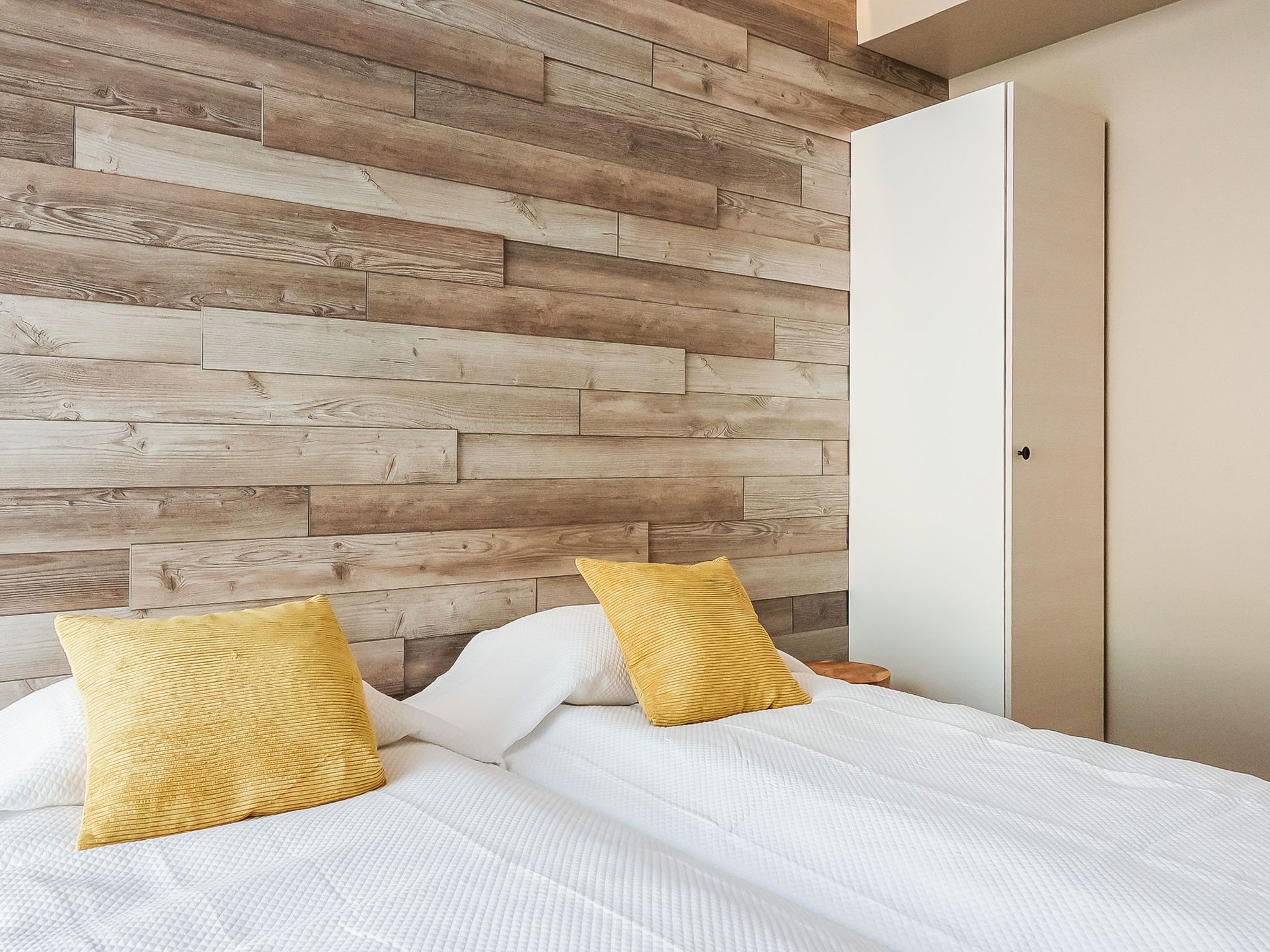A bedroom with a wooden wall panel design, a bed with white bedding and two yellow pillows, and a closed door.