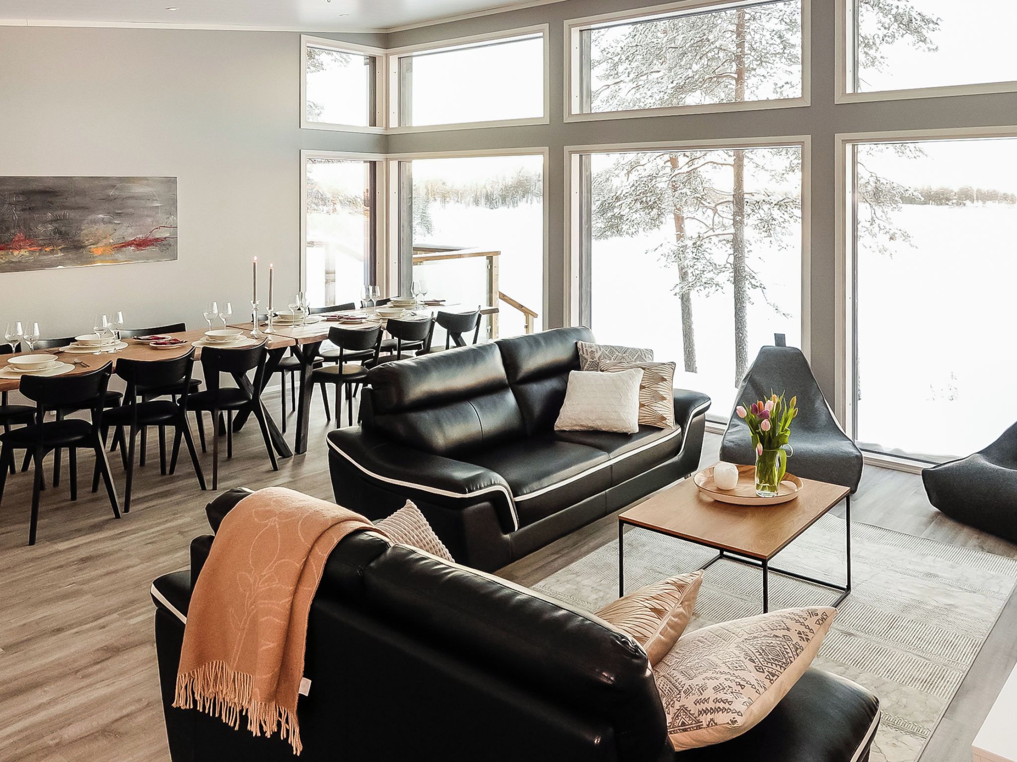 A modern living room with a black leather sofa, a dining area with a wooden table, and large windows offering a view of trees.