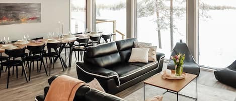 A modern living room with a black leather sofa, a dining area with a wooden table, and large windows offering a view of trees.