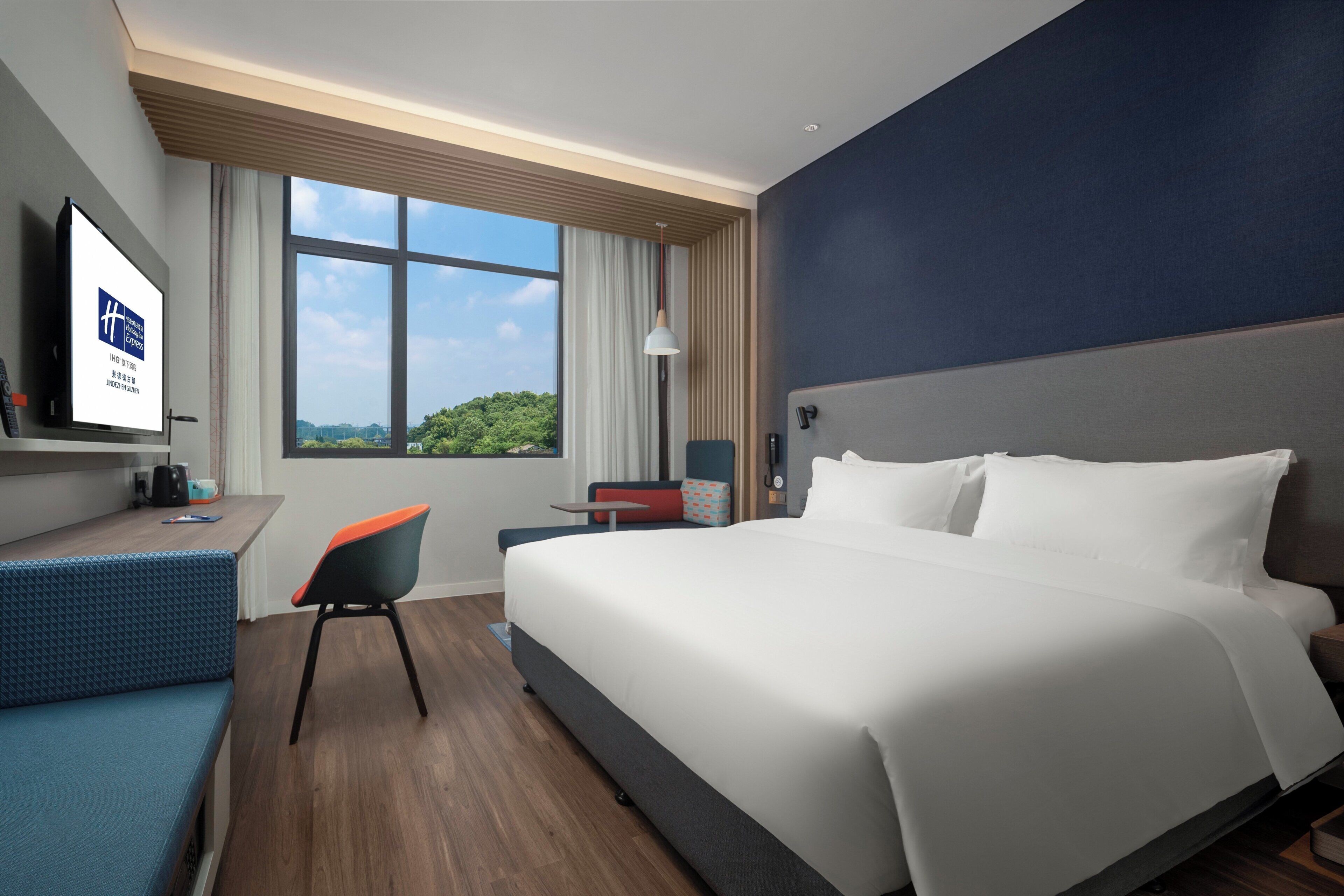 A hotel room with a large bed, a desk with a chair, a television, and a window with a view of greenery.