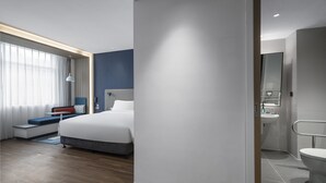 Una camera d'albergo moderna con un letto grande, una zona salotto con una panca e dei cuscini, un comodino e vista sul bagno.