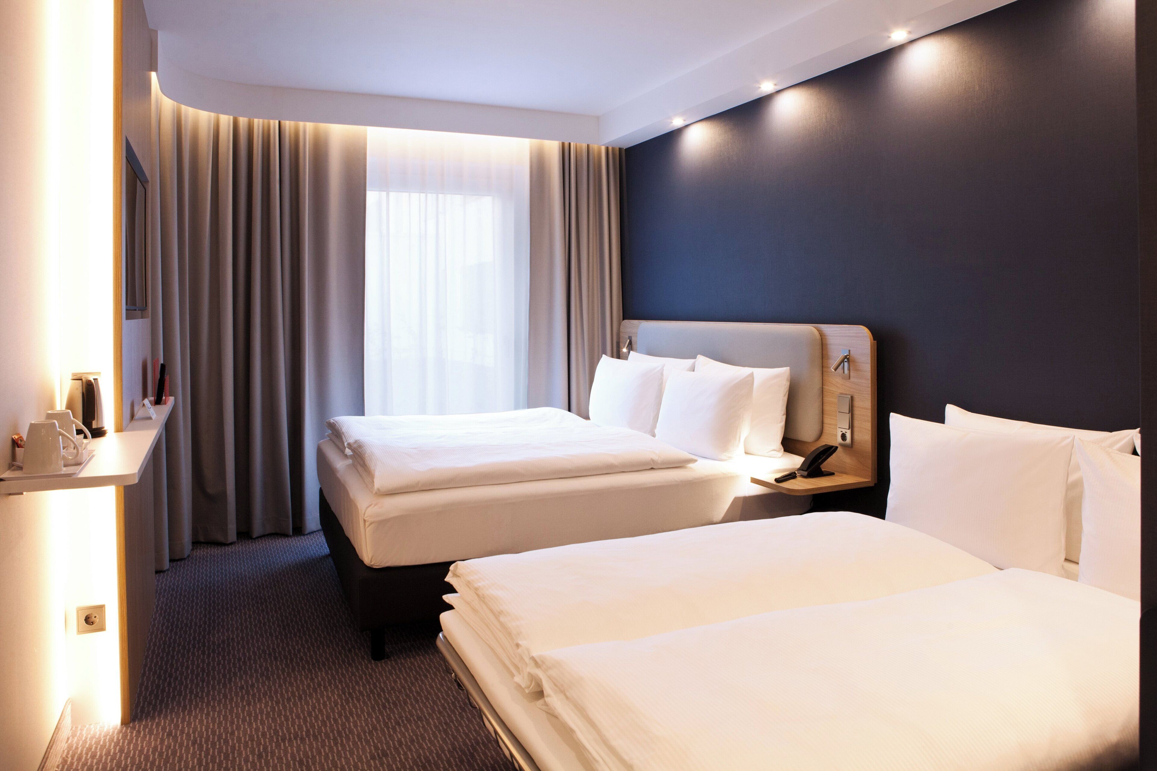 A hotel room with two beds, a bedside table with a phone, and a window with curtains.