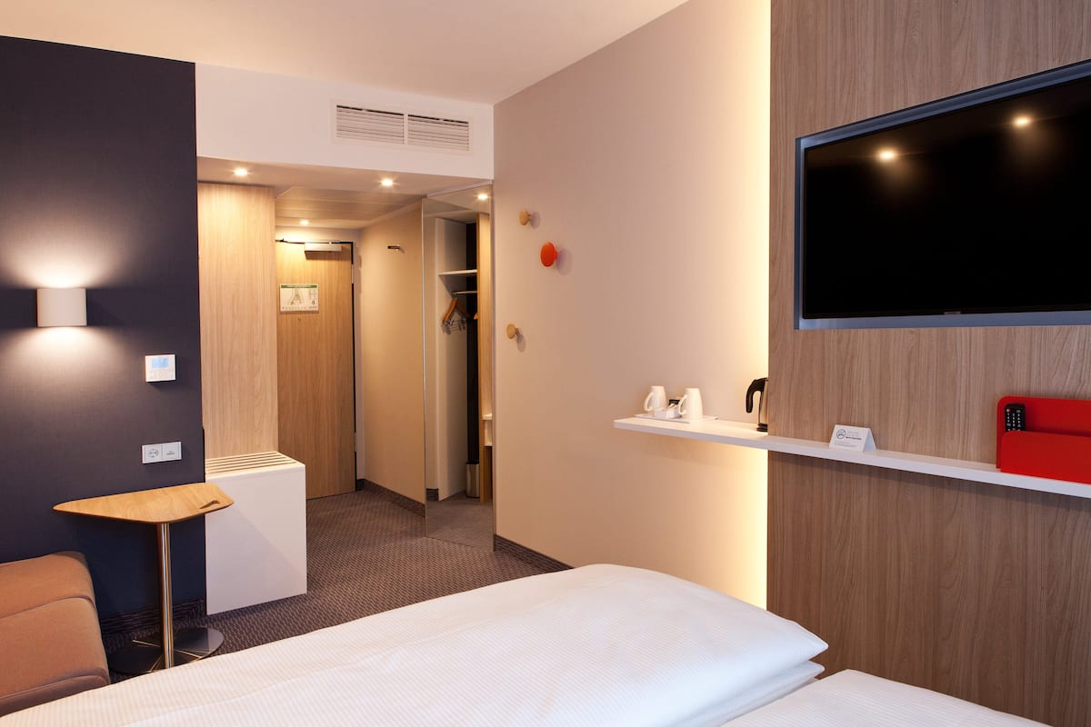 Une chambre d’hôtel avec un lit, une petite table, un miroir, une télévision et une étagère sur laquelle se trouvent une bouilloire et un téléphone rouge.